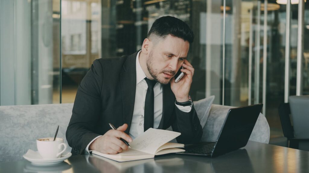 Businessman talks on phone while writing notes in a modern cafe.